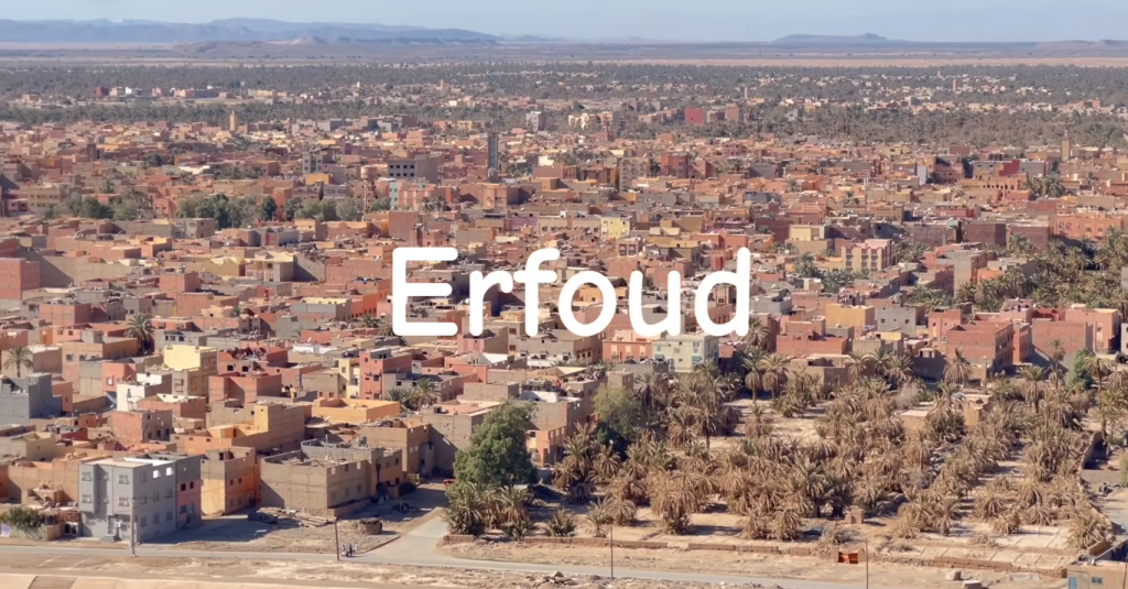 "An aerial view of Erfoud, Morocco, showcasing its picturesque desert landscape with golden dunes stretching toward the horizon, dotted with palm trees and traditional buildings, highlighting its charm as the gateway to the Sahara Desert."