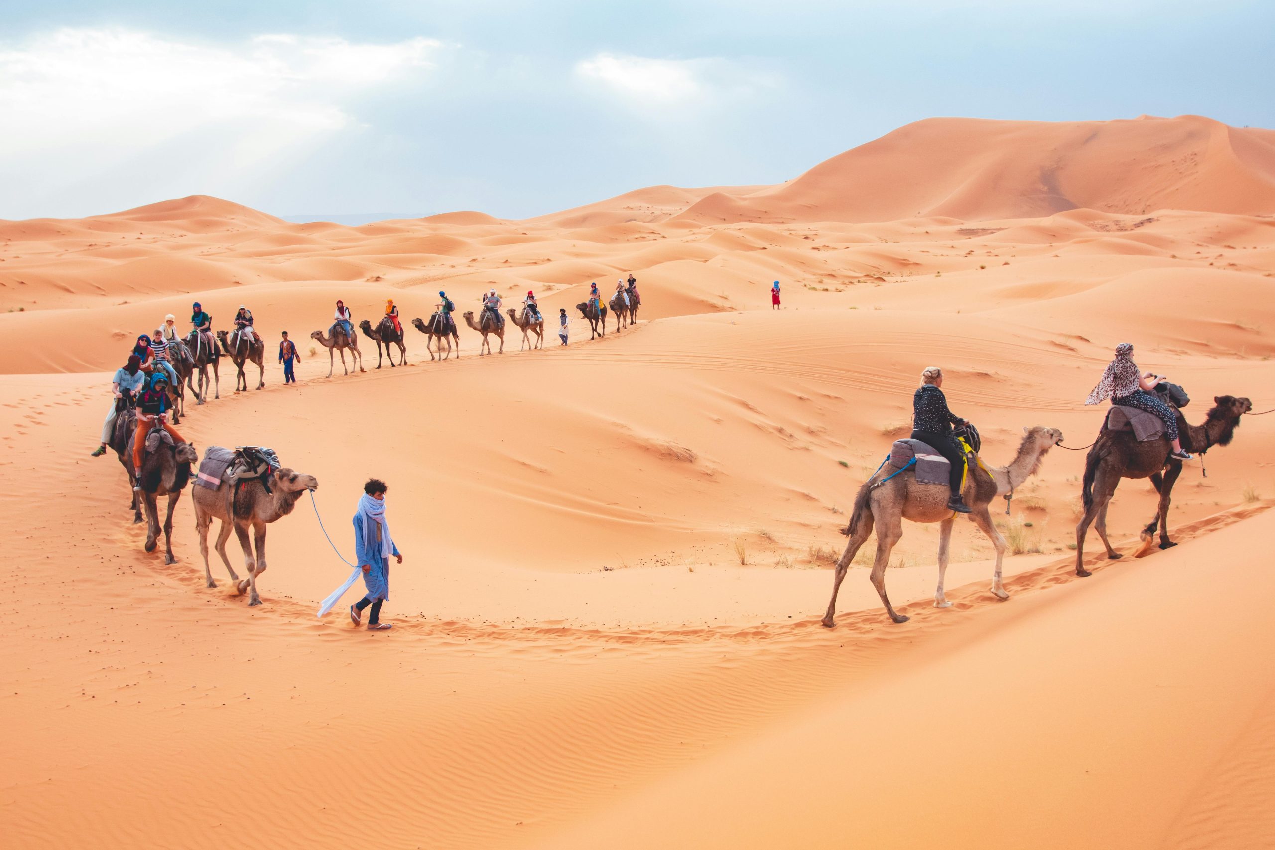 "Golden desert dunes under a vibrant sunset, a line of camels led by a guide in traditional Moroccan attire, with a backdrop of rugged mountains. Experience the enchantment of a Moroccan desert safari."